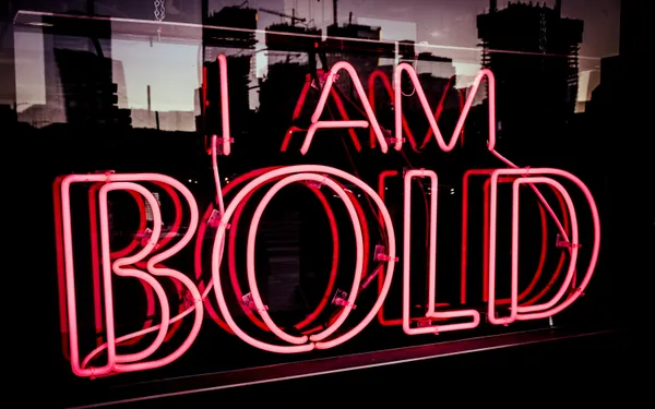 A pink neon sign reading 'I Am Bold' against a dark cityscape backdrop